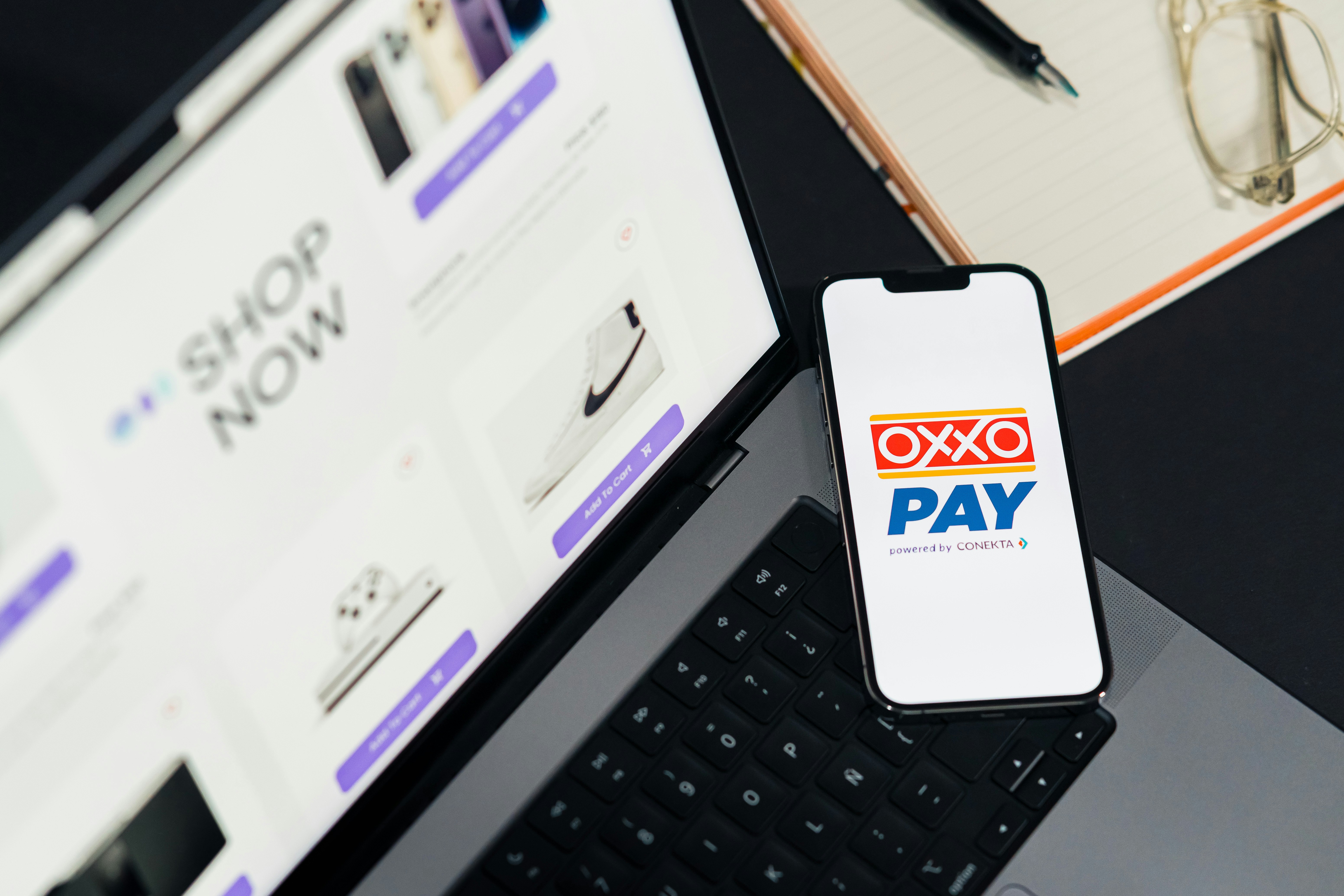 OXXO Pay on phone screen. Top view of workspace. Table desk with laptop with ecommerce on screen, mobile, notepad